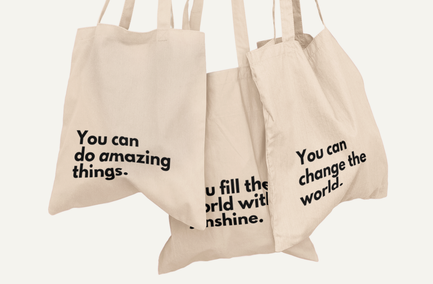 Three natural coloured Affirmation Effect tote bags hanging on a white wall. The tote bags have the words 'You can do amazing things', You fill the world with sunshine', and 'You can change the world' printed in black on the front.