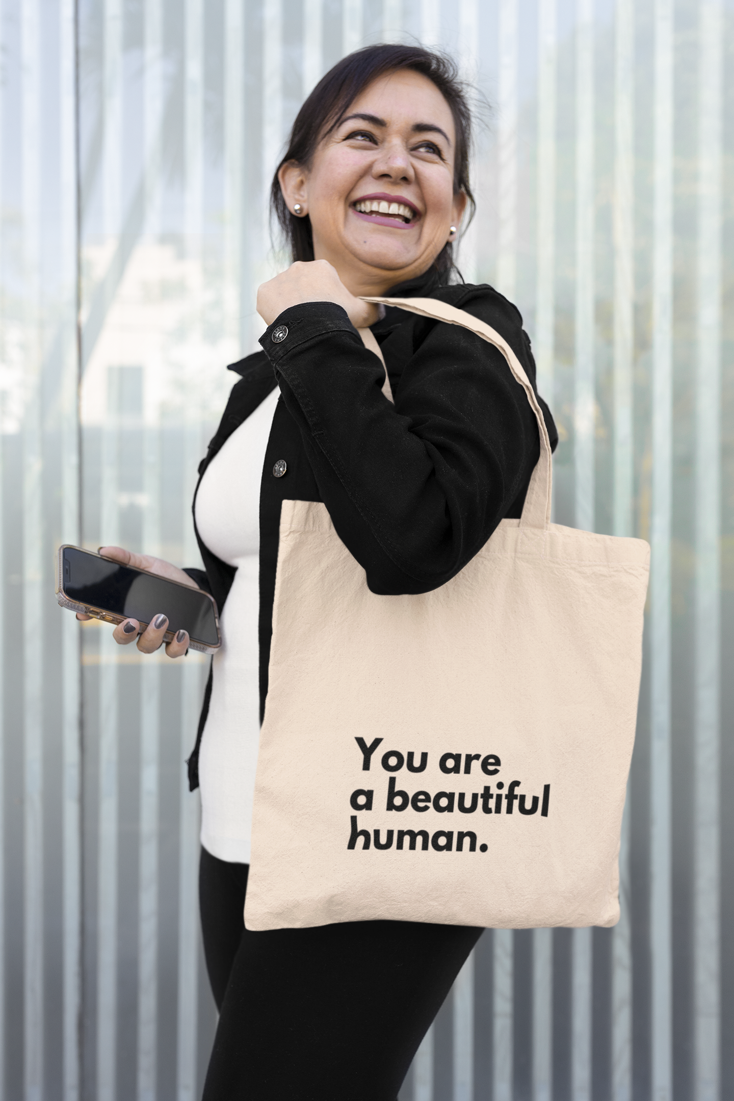 Young women standing in front of corrugated iron, looking back smiling and holding a mobile phone and carrying a natural coloured Affirmation Effect tote bag that says "You are a beautiful human.'