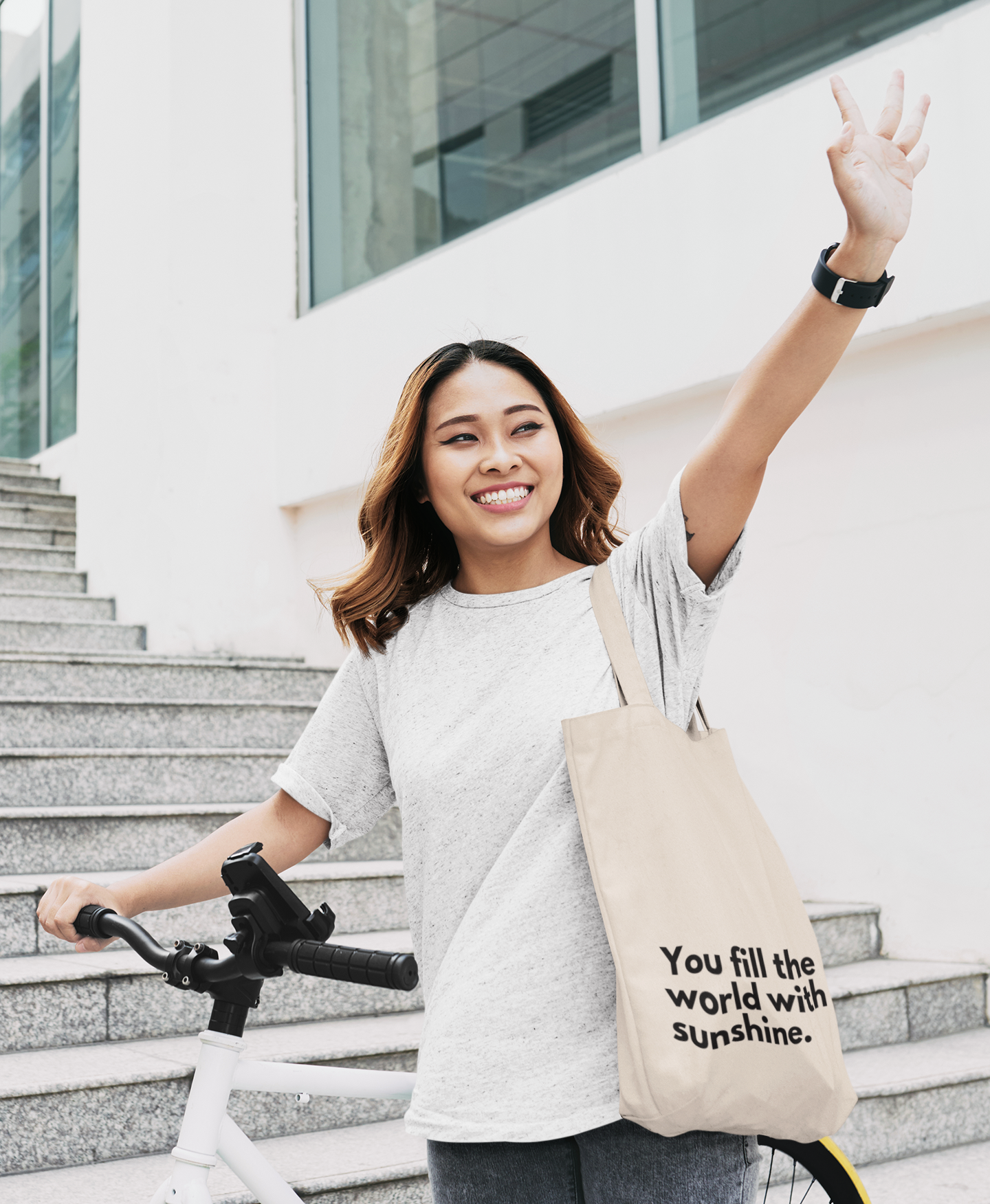 Young women holding a bike and waving. She is carrying a natural coloured Affirmation Effect tote bag with the words 'You fill the world with sunshine' printed in black on the front.