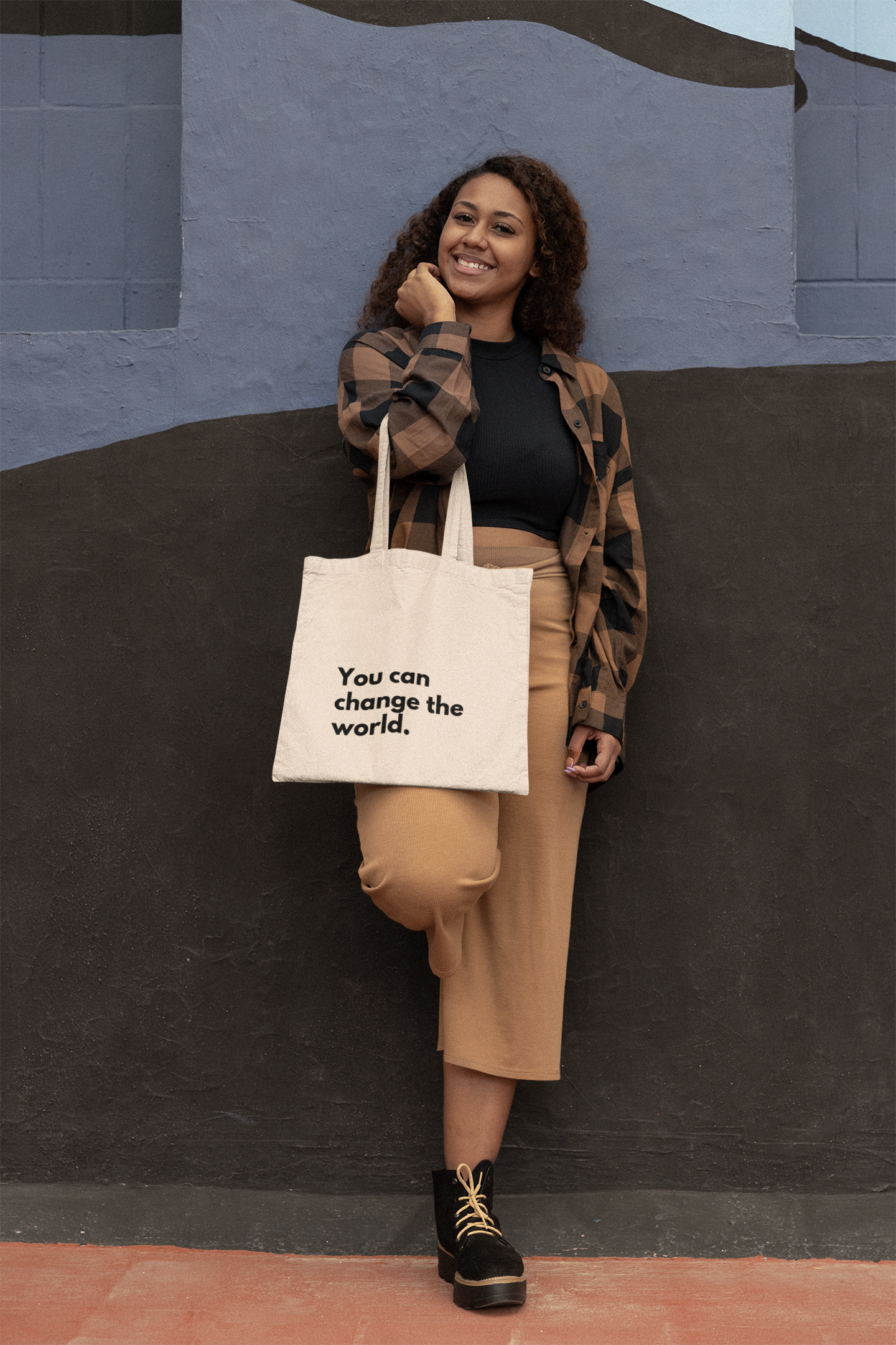 Young women smiling leaning against a painted brick wall. She is carrying a natural coloured Affirmation Effect tote bag with the words 'You can change the world' printed on the front in black.