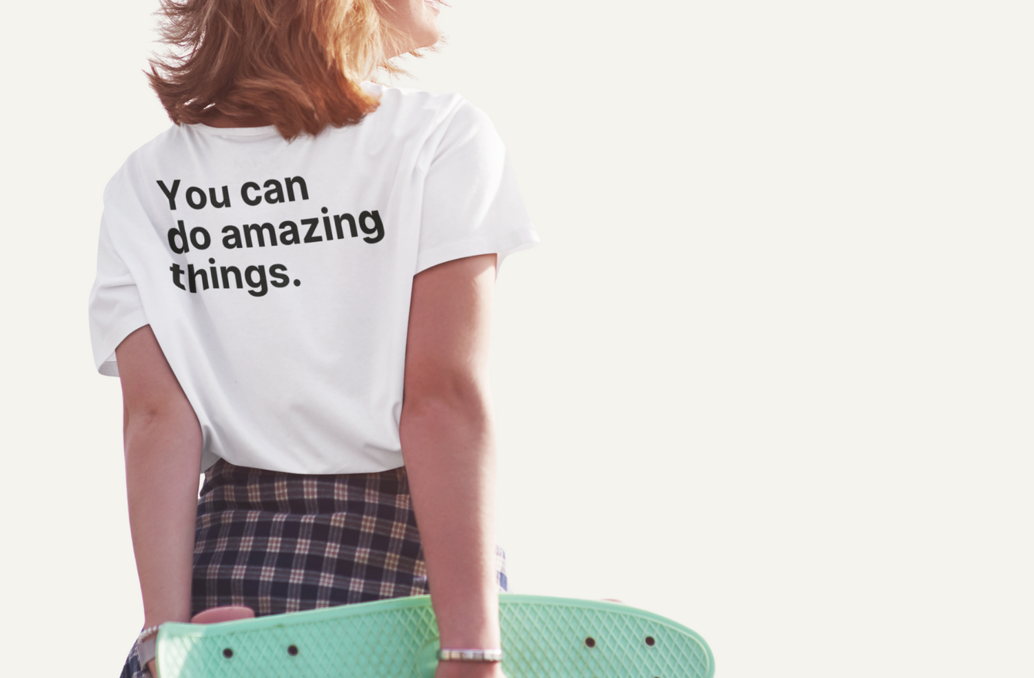 Young women looking off into the distance and holding a green skateboard behind her book. She is wearing a white Affirmation Effect T-shirt with the words 'You can do amazing things' printed in black on the back.